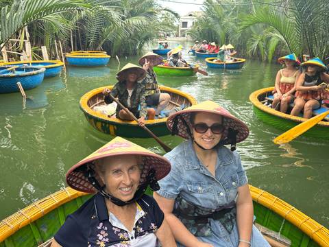 Tourists wearing conical hats smile from round basket boats on a narrow green waterway lined with palms.