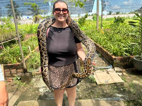 Traveller smiles while holding a large python draped around her shoulders in a lush garden.