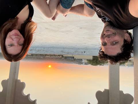 Couple relaxes on a terrace pavilion watching an orange sunset over an Indian cityscape.