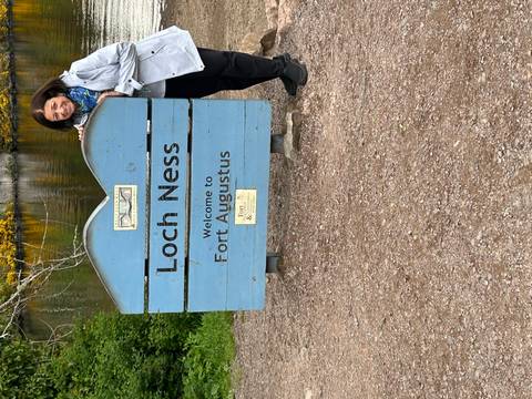A smiling visitor stands beside a blue wooden sign welcoming travelers to Loch Ness at Fort Augustus.