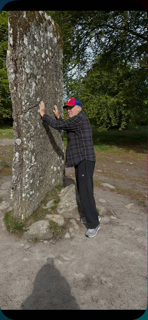 A traveler playfully hugs an ancient standing stone in a wooded clearing.