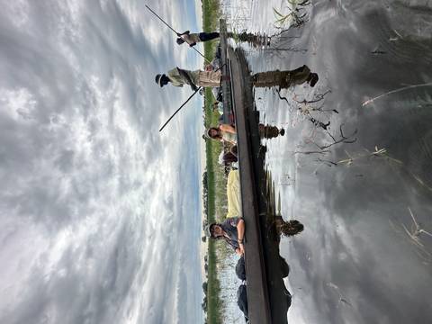 Tourists reclining in a dug-out canoe while a guide poles along a reflective wetland under dramatic cloudy skies.