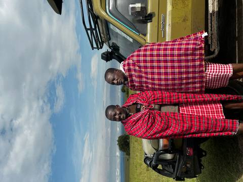 Two Maasai men wrapped in bright red shuka blankets standing beside a safari jeep under a partly cloudy sky.