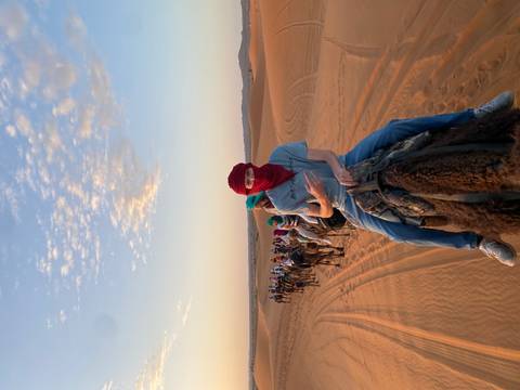 A long caravan of camel riders snakes across glowing orange dunes at sunrise beneath a pastel sky.
