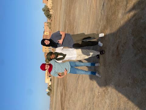 Three friends wearing colourful desert scarves stand smiling on a wide sandy plain with adobe buildings behind.