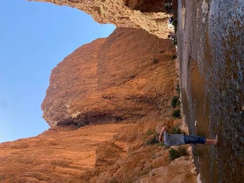 A traveller cools off ankle-deep in a shallow stream at the base of towering red rock canyon walls.