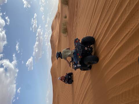 Two riders speed across rolling desert dunes on quad bikes beneath a sky of scattered clouds.