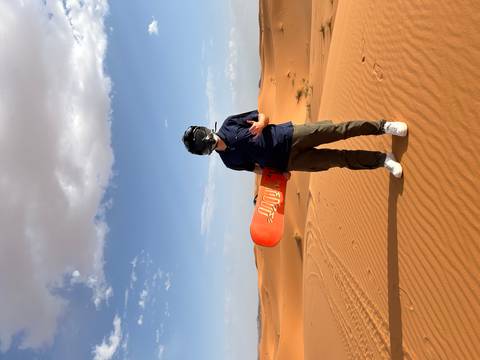 Traveler stands proudly on orange dunes holding a sandboard, helmet on against a bright blue sky.