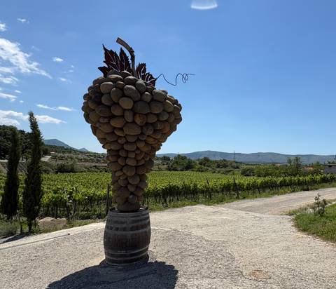Creative sculpture of a grape bunch made of stones atop a barrel in a sunny vineyard landscape.