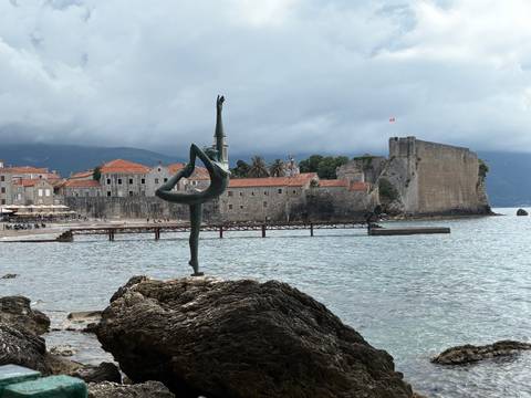 Graceful dancer statue on a rock with Budva Old Town walls and towers across a calm bay.