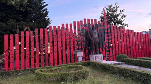 Red-and-black commemorative fence with Albanian flag motif and a statue of a national hero at dusk.