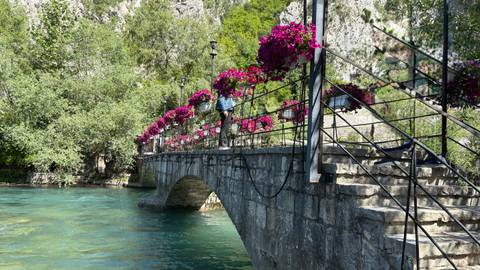 Stone footbridge adorned with vibrant magenta flowers crossing a clear blue river.
