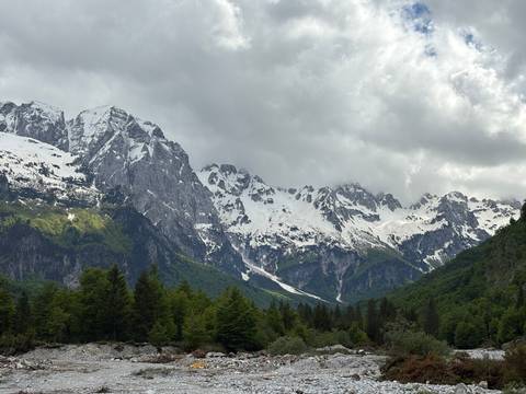 Snow-capped peaks of the Albanian Alps towering over dense green forest under dramatic clouds.