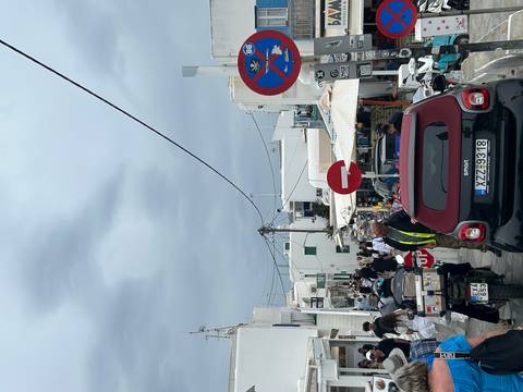 Busy street in Mykonos town filled with scooters, cars, pedestrians and white Cycladic buildings under an overcast sky.