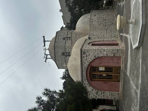 Small stone church with domes and timber doors beneath cloudy skies in Santorini.