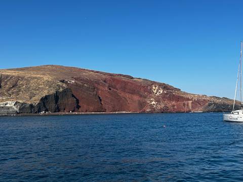 Dramatic red and black volcanic cliffs of Santorini's Red Beach rising from the Aegean Sea with a sailing boat nearby.