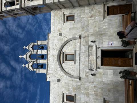 White marble church façade with triple bell tower against a vivid blue sky, visitors at entrance.