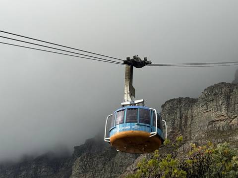 Blue Table Mountain cable car ascends through mist beside sheer rock cliffs under a grey sky.
