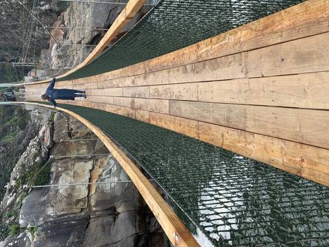 Traveller carefully walks across a long wooden suspension bridge above churning water in a forested gorge.