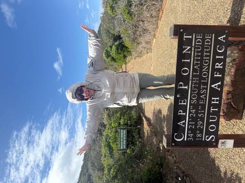 Woman in windbreaker spreads her arms behind the Cape Point latitude and longitude sign under a bright blue sky.