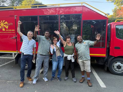 Tour group waves beside a bright red overland safari truck parked in a lot with trees behind.