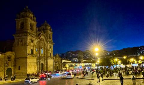 Nighttime scene of a lively colonial square in front of an illuminated cathedral, busy with cars and pedestrians.