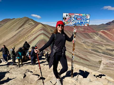 Hiker posing with a colourful sign at the summit of Rainbow Mountain with multicoloured ridges behind.