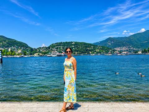 Woman in floral dress standing in clear blue lake with green hills and boats behind her.