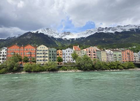 Row of colourful riverside houses with snowy Alpine mountains towering behind, reflected in turquoise water.