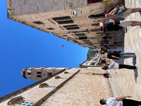 Crowded limestone street inside Dubrovnik Old Town with bell tower and dove flying overhead.