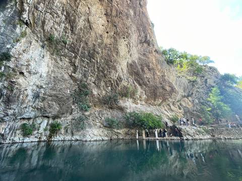 Steep rocky cliff towering above emerald spring where visitors explore a narrow path.