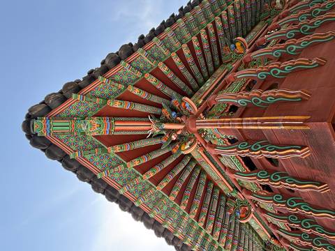 Intricately painted wooden eaves of a Korean temple captured from below against blue sky.