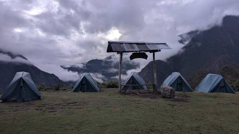Row of green trekking tents pitched on a grassy ridge with misty cloud-filled valleys and dark mountains behind.