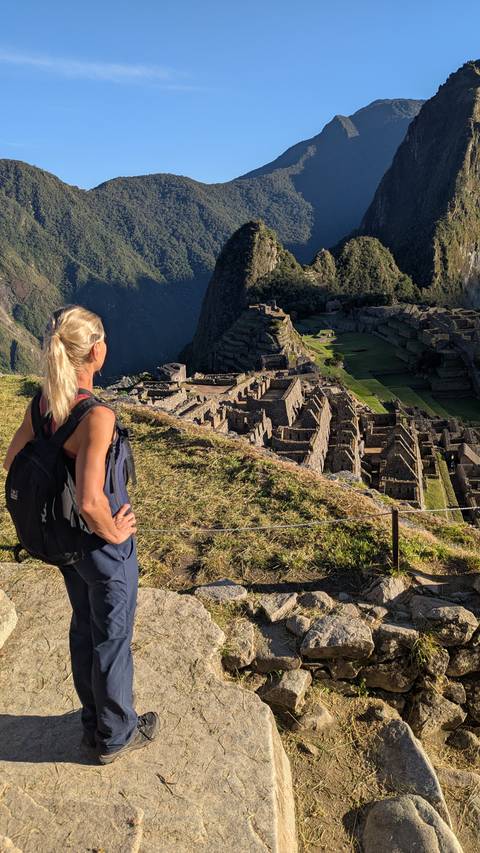 Female trekker gazes over the stone terraces and ruins of Machu Picchu from a grassy lookout.