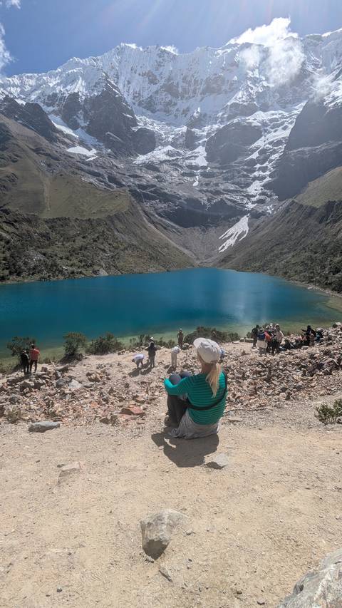 Vivid turquoise glacial lake ringed by steep rocky peaks with trekkers gathered along the stony shore.