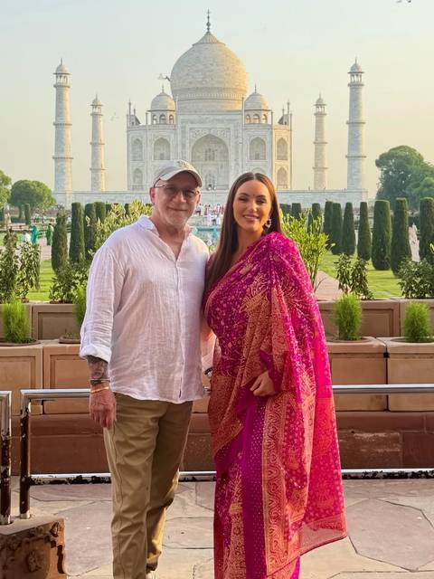 Couple dressed smartly pose in front of manicured gardens with the Taj Mahal gateway in the distance.