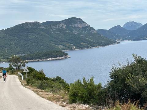 Cyclist descends quiet coastal road overlooking deep blue Adriatic coves and forested hills.