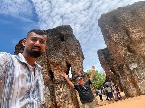 Guide and traveler pose among tall red-brick ruins of an ancient Sri Lankan city under a patterned sky.