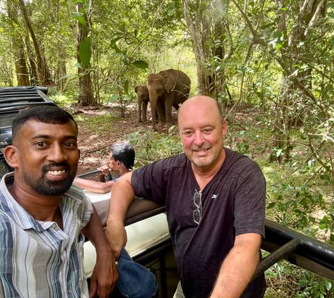 Tourist and guide smile from an open jeep while wild elephants graze in the forest behind them.