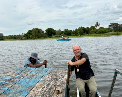 Traveler climbs from a rustic wooden raft while another man rows on a tranquil lake with palm trees beyond.