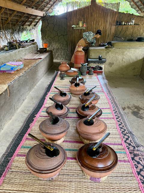 Long table displays rows of traditional clay pots with ladles inside a rustic cooking pavilion.