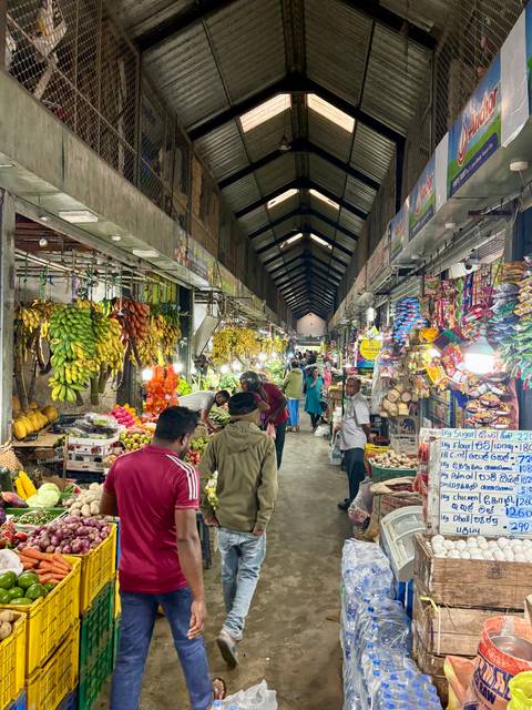 Busy indoor produce market with colourful hanging bananas and fruit stalls as shoppers browse the aisles