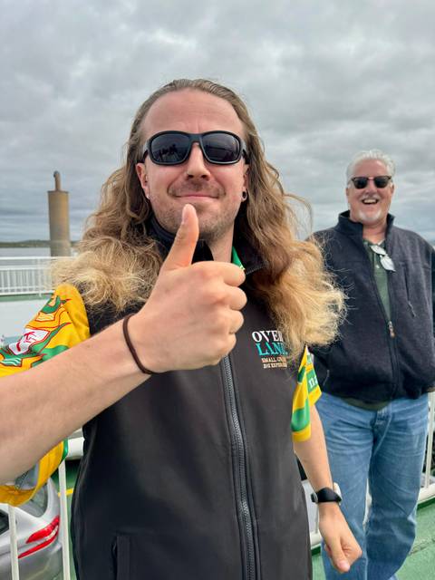 Close-up of a smiling traveler giving a thumbs-up on a ferry deck, with another man laughing in the background against a cloudy sky and distant shoreline.