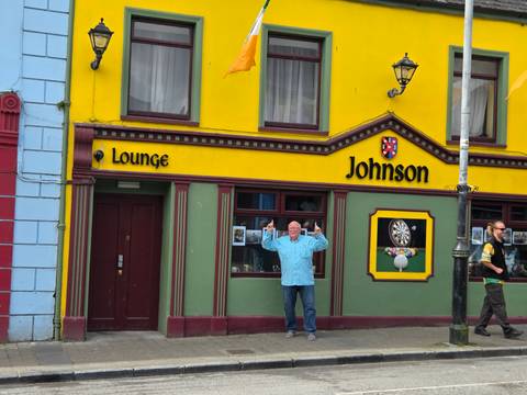 Colorful pub facade with bright yellow walls and a man posing triumphantly in front while another passer-by walks past.
