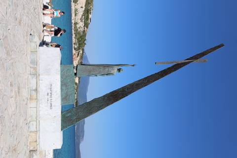 Bronze statue of Pythagoras pointing skyward by a marina with visitors sitting nearby and turquoise sea behind.