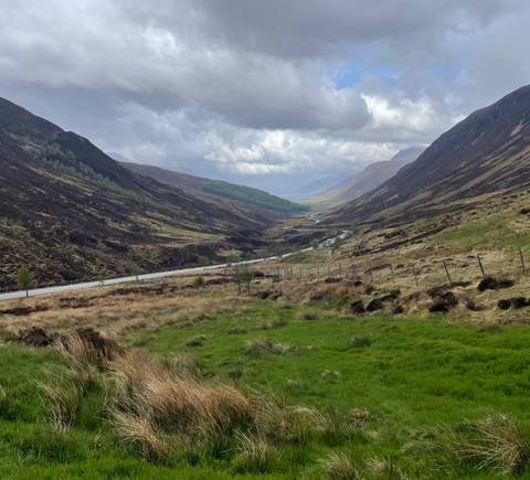 Long Highland valley carpeted with heather and crossed by a winding road under dramatic skies.