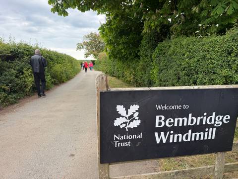 National Trust sign for Bembridge Windmill next to a hedge-lined footpath with walkers.