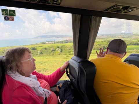 Two passengers inside a coach looking out over green coastal countryside through large windows.