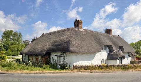 Quaint whitewashed thatched cottage along a rural roadside beneath a bright sky.