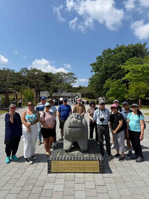 A tour group gathers around a stone monument at an outdoor historical site on a sunny day.